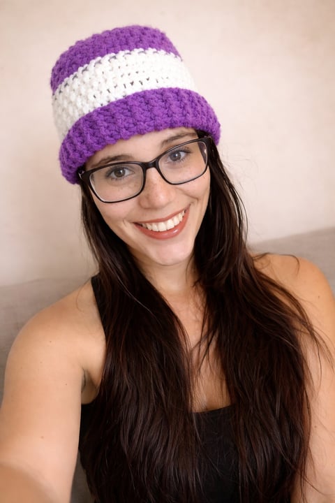 Woman with long dark hair and glasses wearing a purple and white striped crocheted beanie, smiling at the camera