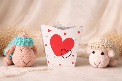 Two knitted toy sheep with a heart-patterned gift bag between them on a beige textured background