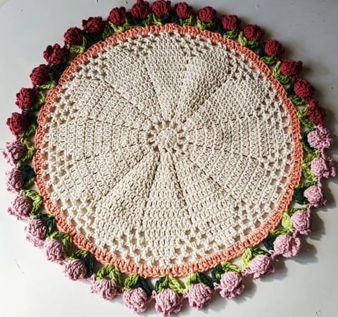 Circular crocheted doily with cream center, coral border, and decorative flower motifs in pink, red, and green around the edge